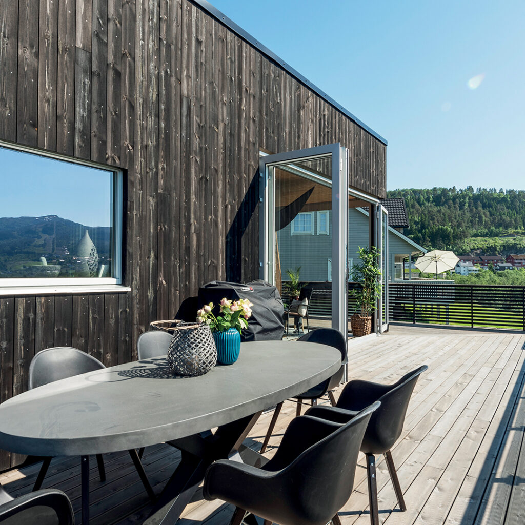 Husvegg med royalimpregnert stående kledning fra MøreRoyal. På terrassen står det et spisebord med stoler og en tildekket gassgrill. I bakgrunnen synes nabolag og grønne skoger.