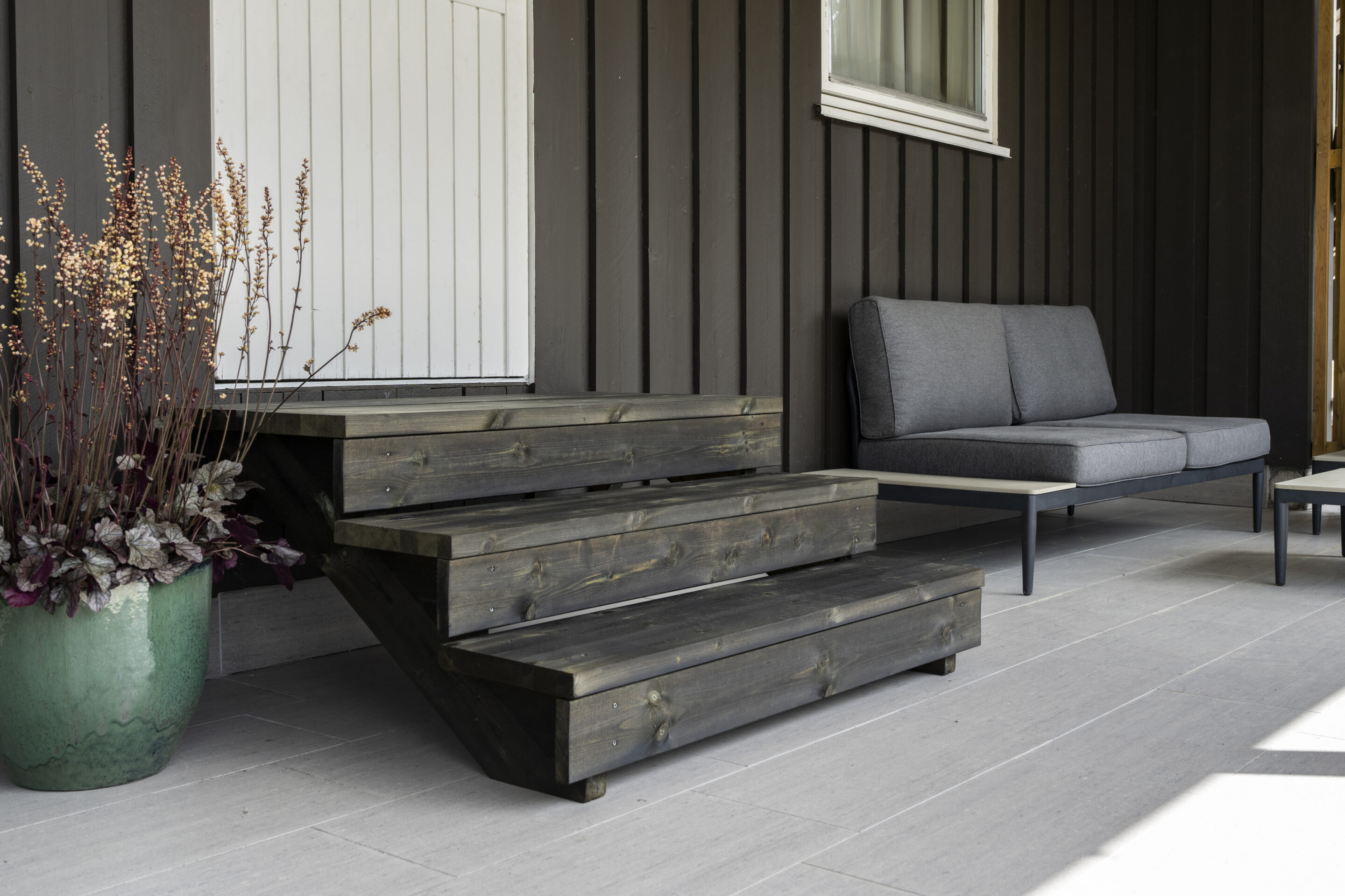 Foto utetrapp med tre trinn, CU-impregnert og oljet svart. En del av Talgøs kjapptrapp-konsept. Ferdig montert trapp opp til verandadør. Grønn krukke med plante til venstre for trappen. Utesofa med grå puter til høyre for trappen.